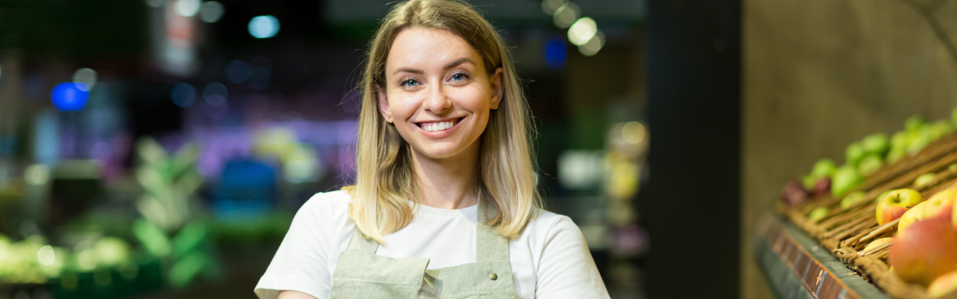 Junge Mitarbeiterin im Supermarkt steht lächelnd mit verschränkten Armen in der Obst- und Gemüseabteilung.